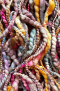 A close-up pile of chunky, twisted yarn in a variegated mix of pastel pink, mustard yellow, lavender, gray, and brown tones. Hand-dyed and hand spun in Minneapolis, merino wool.