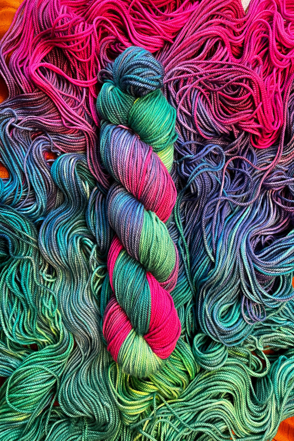 One twisted skein of Bohemian Rhapsody hand dyed yarn in greens, blues, purples and shocking magenta on a flat skein of the same yarn. 