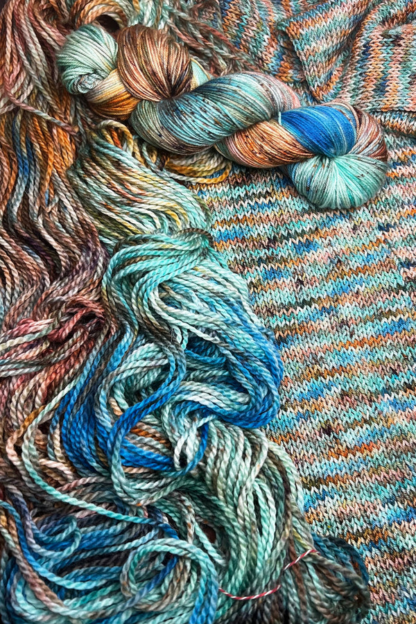 Dancer in Green hand-dyed yarn display with skeins of yarn and knit sample. Hand dyed in Minneapolis by Painted Skein with sage green, peach, sapphire tones and with speckles of brown.