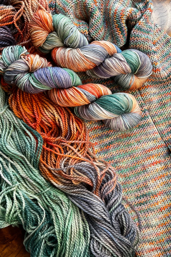 Sunrise yarn display with skeins of yarn and knit sample. Hand dyed in Minneapolis by Painted Skein with greens, lavender and peachy orange tones.