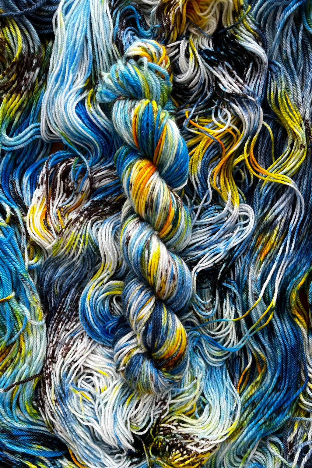 One twisted skein of colorful of hand-dyed Starry Night yarn with blue, yellow, orange and white hues, on top of flat skein in same colorway. 